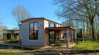 Foto van een moderne tiny house met houten gevel en grote ramen, omringd door bomen en zonlicht.