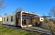 Moderne Modus-lodge in Bad Hoophuizen, Nederland, met houten terras en tuinmeubilair onder een blauwe lucht.