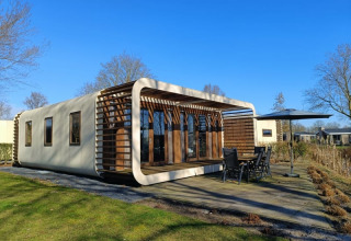 Moderne Modus-lodge bij Bad Hoophuizen in Nederland, met houten terras en tuinset onder blauwe lucht.
