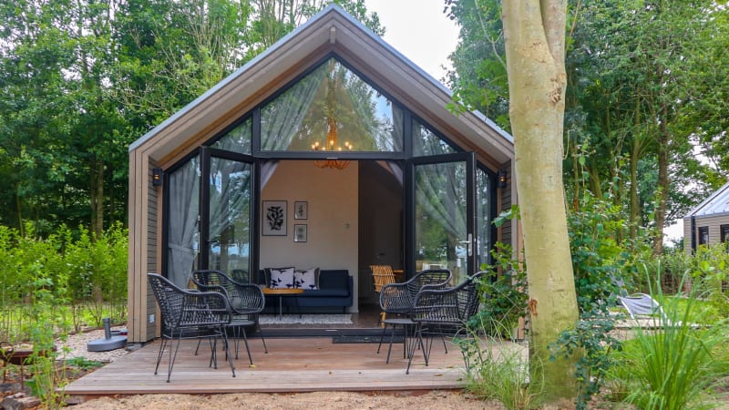 Beach House Lodge mit moderner Glasfront, Terrasse und Gartenmöbeln, umgeben von grüner Natur und Bäumen.