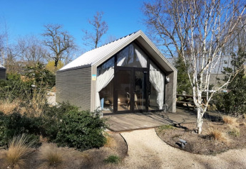 Et lille strandhus-lodge med moderne design, omgivet af natur og træer under en blå himmel.