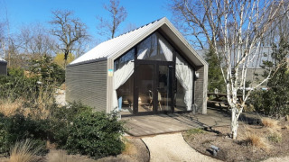 Et lille strandhus-lodge med moderne design, omgivet af natur og træer under en blå himmel.