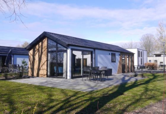 Moderne lodge ved Velthorst med store vinduer, terrassemøbler og grønne omgivelser under blå himmel.