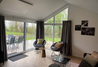Modernes Wohnzimmer in einer Velthorst-Lodge mit großen Fenstern, Blick auf Terrasse und Garten in Bad Hoophuizen.
