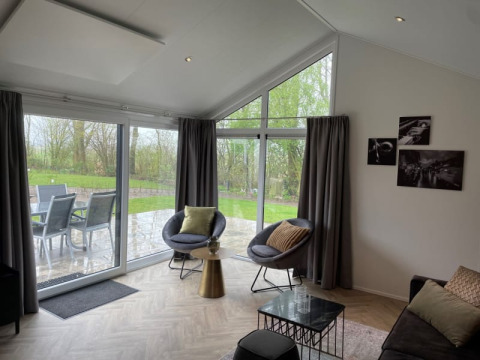 Moderne lodge-stue med store vinduer, runde stole, udsigt til terrasse og grøn have ved Bad Hoophuizen.