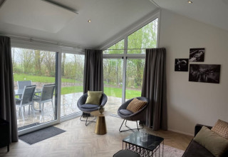 Moderne lodge-stue med store vinduer, runde stole, udsigt til terrasse og grøn have ved Bad Hoophuizen.
