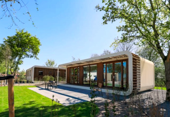 Moderne Modus Wellness-lodge med glasfacade midt i naturen, omgivet af grønne træer og græsplæne.