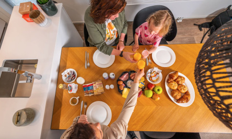 Een gezin geniet van een ontbijt met broodjes, fruit en drankjes aan een houten tafel in een glamping accommodatie.