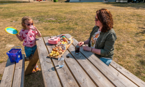 Moeder en dochter genieten van een picknick aan een houten tafel op een grasveld in een vakantiepark.
