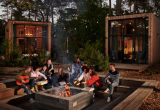 Groep vrienden geniet bij een kampvuur voor moderne glampinghutten in een sfeervol vakantiepark.