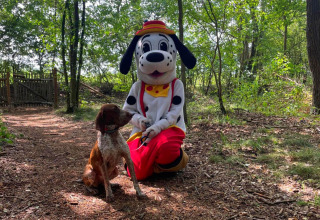 Persoon in een hondenmascottekostuum en een echte hond poseren samen in een bos bij Papillon Country Resort.