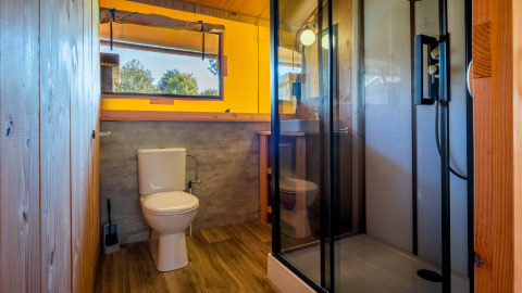 Baño moderno en una cabaña con paredes de madera, ducha, inodoro y una amplia ventana con vista verde.