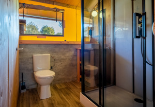 Baño moderno en una cabaña con paredes de madera, ducha, inodoro y una amplia ventana con vista verde.