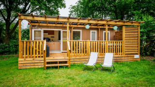 Die Cottage Next XL Holzhütte mit überdachter Terrasse und zwei Stühlen im grünen Gartenbereich.