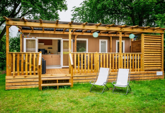 Cabaña de madera Cottage Next XL con terraza cubierta y dos sillas de jardín en entorno verde y frondoso.