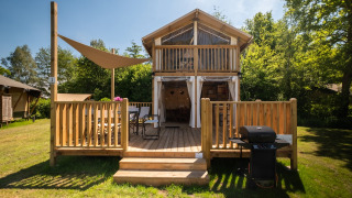 Air Lodge 2.0 met houten veranda, zithoek en barbecue in een groene omgeving met veel bomen.