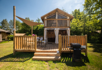 Air Lodge 2.0 met houten veranda, zithoek en barbecue in een groene omgeving met veel bomen.