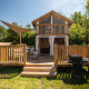 Air Lodge 2.0 med træterrasse, udendørs spisebord og grill, omgivet af grønne træer og solskin.
