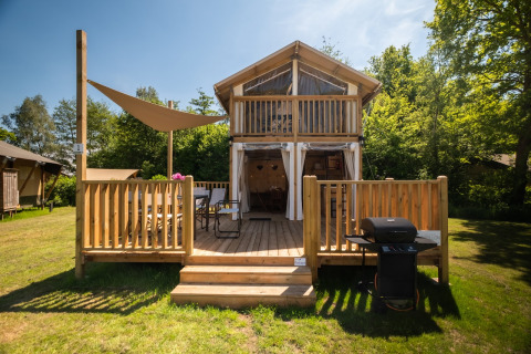 Air Lodge 2.0 met houten veranda, zithoek en barbecue in een groene omgeving met veel bomen.