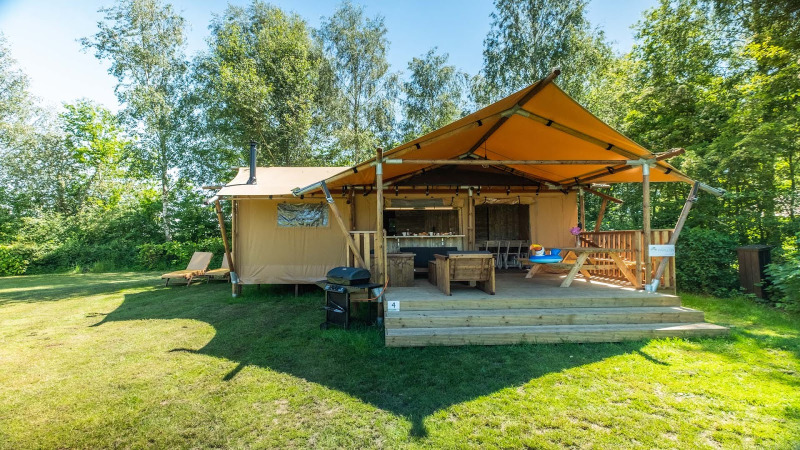 Imagen soleada de Safarilodge Deluxe con terraza cubierta, barbacoa y zonas verdes rodeadas de árboles.
