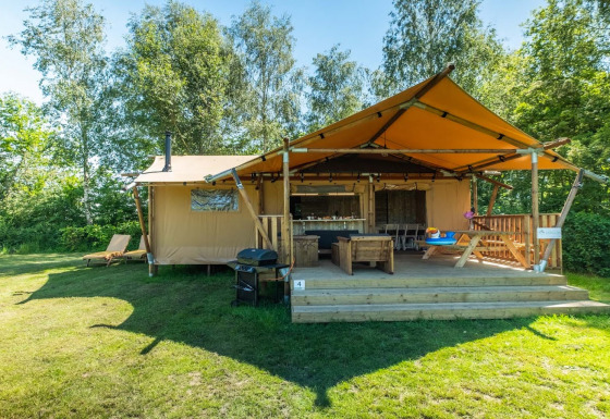 Safarilodge Deluxe in het zonlicht met een overdekt terras, barbecue en groene graszone tussen bomen.