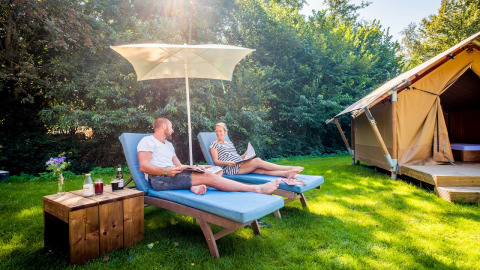 Zwei Personen entspannen auf Liegestühlen unter einem Sonnenschirm vor einem Safarizelt im Papillon Country Resort.
