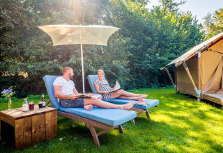 Zwei Personen entspannen auf Liegestühlen unter einem Sonnenschirm vor einem Safarizelt im Papillon Country Resort.