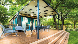 Freed Home lodge met een groot houten terras, buitenmeubels en blauwe gordijnen omgeven door bomen.
