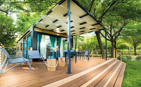 Freed Home lodge with a spacious wooden deck, outdoor seating, and blue curtains surrounded by lush trees.