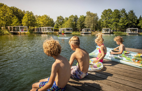 Kinderen genieten van de zon op een steiger aan het water, met moderne vakantiehuisjes op de achtergrond.