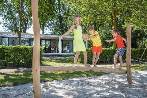 Drie kinderen spelen op een evenwichtsbalk in een speeltuin bij vakantiepark BreeBronne in Limburg.