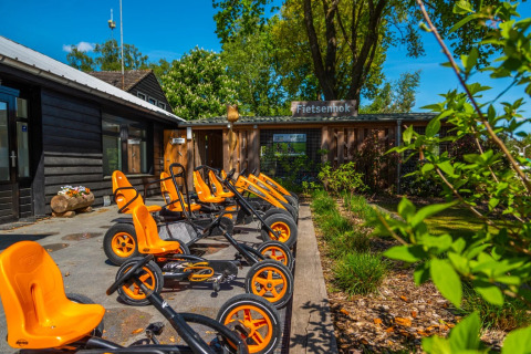 Rij oranje trapkarts geparkeerd voor het Fietsenhok bij een vakantiepark met glamping accommodaties.