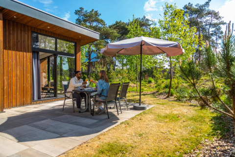 Koppel geniet van een maaltijd buiten onder een parasol bij een houten vakantiehuis te midden van de natuur.