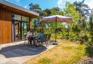 Koppel geniet van een maaltijd buiten onder een parasol bij een houten vakantiehuis te midden van de natuur.