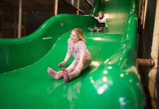 Twee kinderen glijden van een groene glijbaan in een indoor speeltuin bij een vakantiepark met glamping.