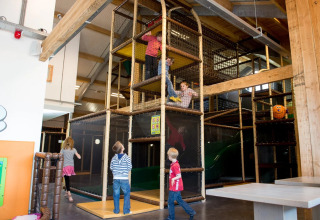 Kinderen spelen in een grote indoor speeltuin met houten en netconstructies in een vakantiepark.