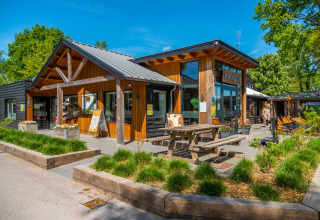 Moderne receptie van een vakantiepark met houten gevel, picknicktafel en groene beplanting onder een blauwe hemel