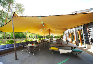 Gezellige glamping zithoek bij een vakantiepark met grote gele overkapping, buitentafels en banken.