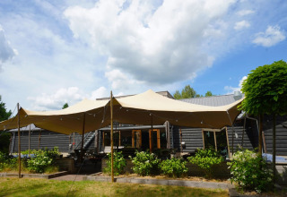 Glampingtent bij een vakantiepark, ruime luifel en houten chalet met groene tuin, zon en wolkenlucht.