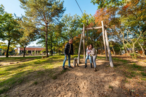 Gezin geniet van een zonnige dag in een vakantiepark met glamping, naast een houten schommel in het bos.