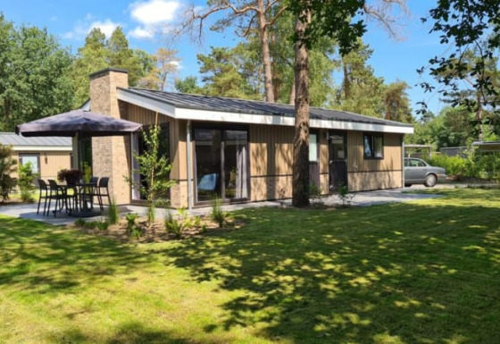 Moderna casa de campo con gran jardín, zona de comedor al aire libre y rodeada de altos árboles verdes.