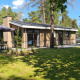 Et moderne lodgehus med stor have, udendørs spiseområde og omgivet af grønne træer under blå himmel.