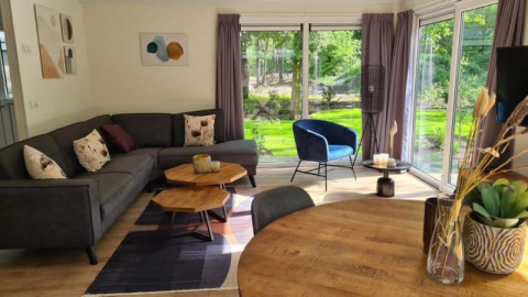 Moderne lodge-stue med gråt hjørnesofa, blå stol, store vinduer, træbord og udsigt til grøn have.