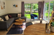 Salon moderne de lodge avec canapé d’angle gris, fauteuil bleu, grandes fenêtres et vue sur un jardin vert.