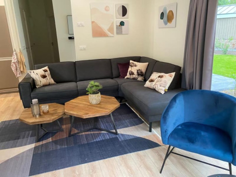 Moderne lodge-stue med mørkegrå hjørnesofa, blå stol, sekskantede borde, planter og abstrakt kunst.