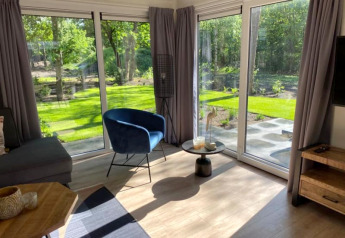 Gezellige lodge woonkamer met grote ramen, grijze gordijnen, blauwe stoel en zicht op de groene tuin.