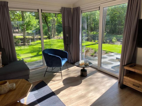 Salon lumineux d’un lodge avec grandes baies vitrées, rideaux gris, fauteuil bleu et vue sur jardin verdoyant.