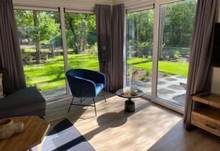 Sala de estar luminosa en una cabaña con grandes ventanales, cortinas grises, sillón azul y vista al jardín.