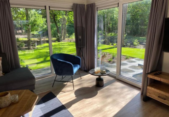 Gezellige lodge woonkamer met grote ramen, grijze gordijnen, blauwe stoel en zicht op de groene tuin.