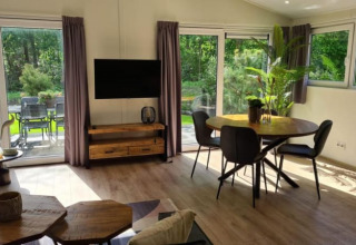 Moderne lodge-stue med runde spisebord, sorte stole, store vinduer, TV, træmøbler og udsigt til grøn have.